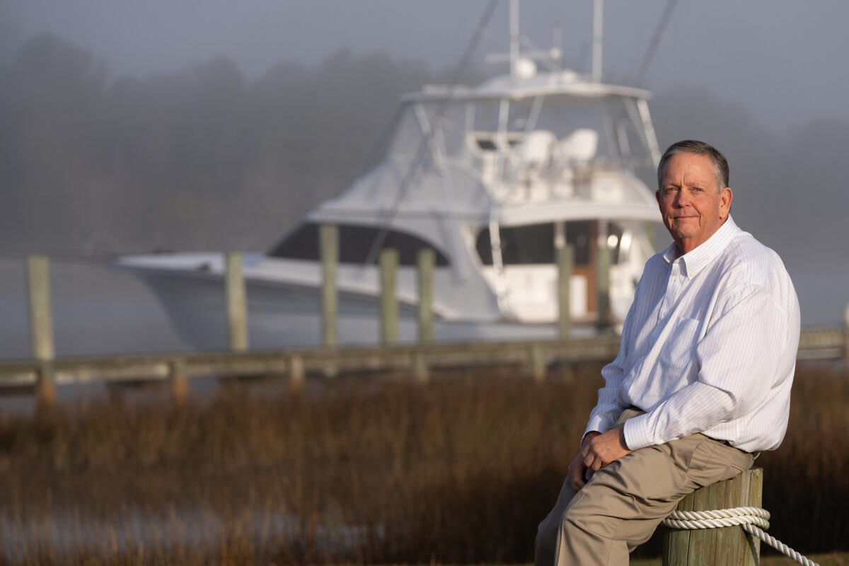 Randy Ramsey, founder of Jarrett Bay Boatworks, seated on a dock piling with a custom sportfishing yacht in the misty background