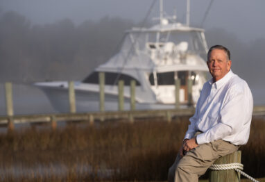 Randy Ramsey, founder of Jarrett Bay Boatworks, seated on a dock piling with a custom sportfishing yacht in the misty background