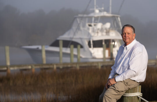 Randy Ramsey, founder of Jarrett Bay Boatworks, seated on a dock piling with a custom sportfishing yacht in the misty background