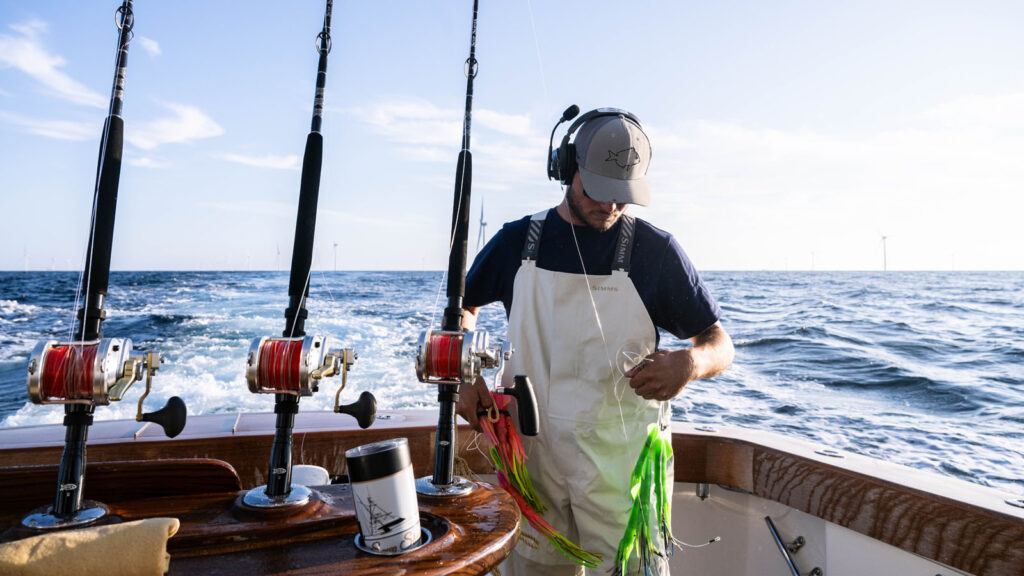 Mate in a white Simms bib holding trolling lures in the cockpit with offshore wind turbines on the horizon