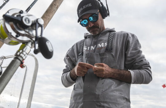 Offshore angler in Simms Rip Line Pro bibs rigging a leader beside rods and reels in the rocket launcher