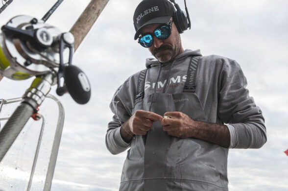 Offshore angler in Simms Rip Line Pro bibs rigging a leader beside rods and reels in the rocket launcher