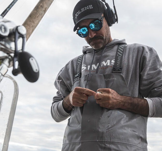 Offshore angler in Simms Rip Line Pro bibs rigging a leader beside rods and reels in the rocket launcher
